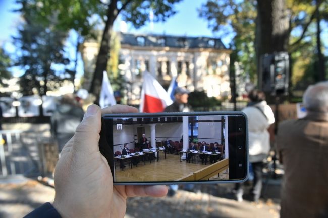 Obrady TK oglądane na ekranie telefonu na tle demonstracji przed siedzibą Trybunału Konstytucyjnego w Warszawie, fot. PAP/Radek Pietruszka